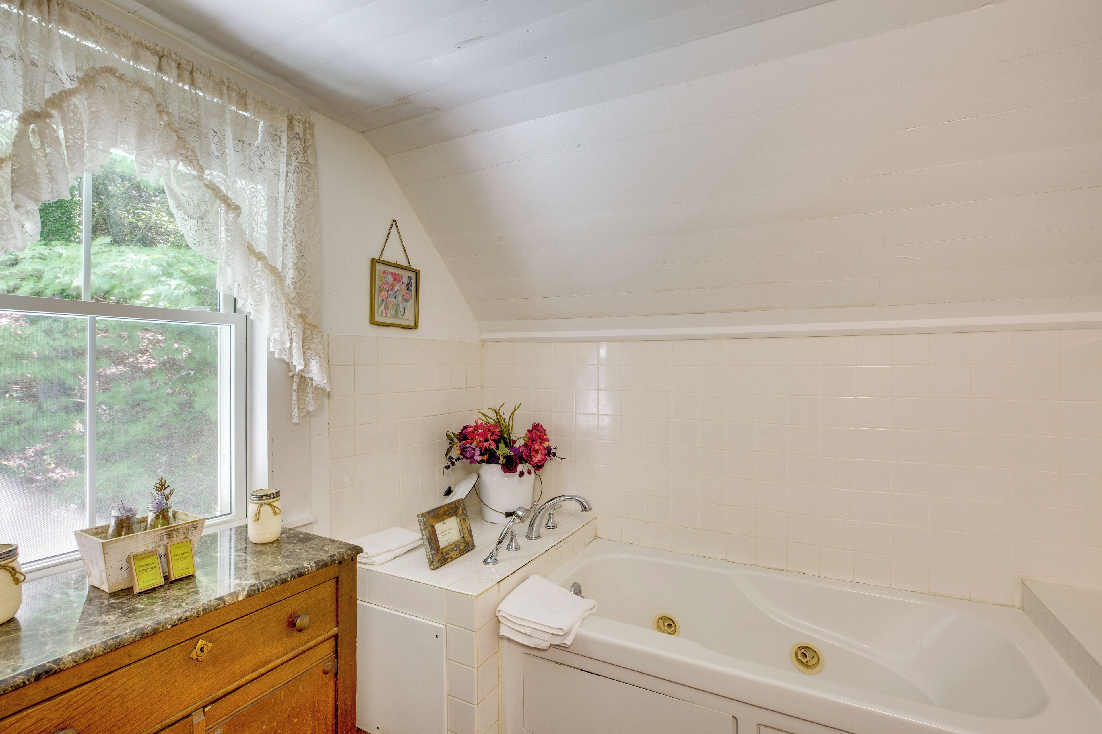 Un baño con bañera, tocador con espejo, ventana con cortinas de encaje y un jarrón con flores en la encimera.