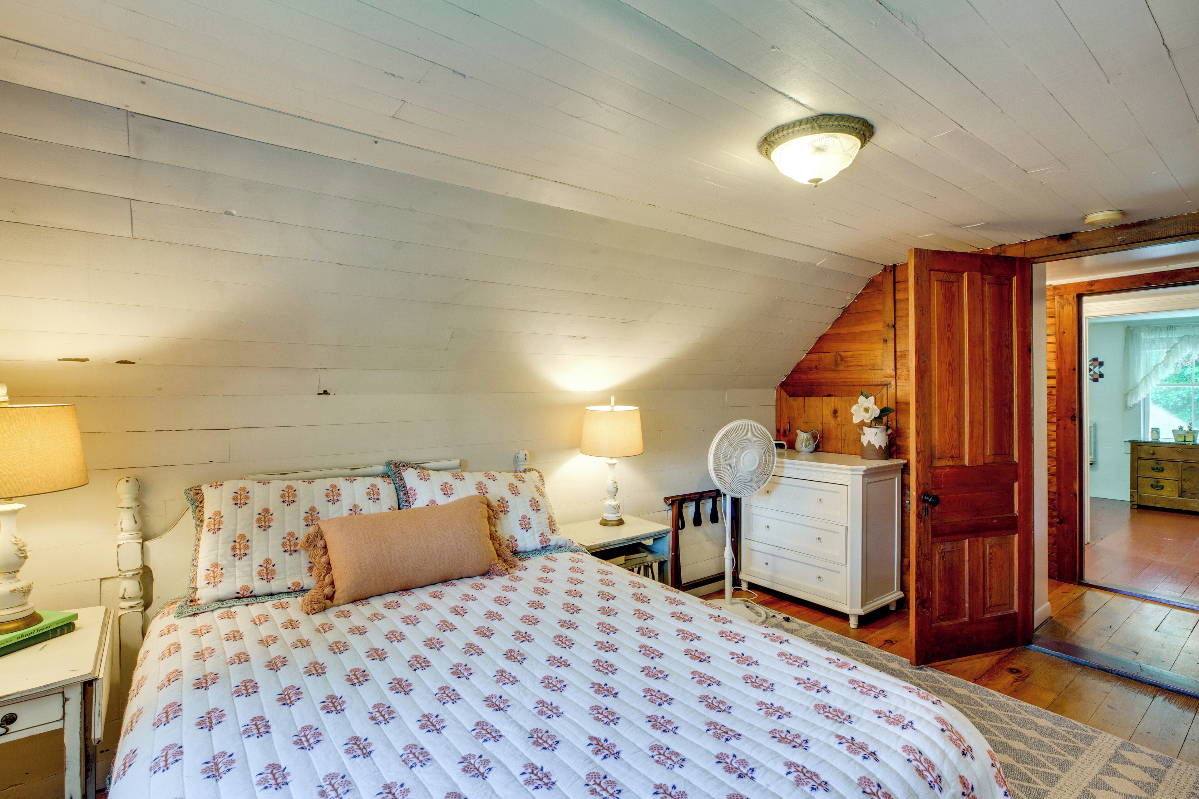A bedroom with a bed, bedside lamps, a dresser, a fan, and a door leading to another room.
