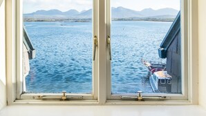 A window with a view of a calm body of water, a dock with a boat, and distant mountains. - Roundstone Quay (Roundstone)