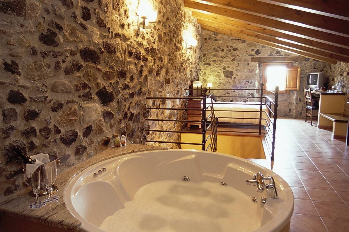 Ein Badezimmer mit Steinsäulen, einer großen Badewanne, Holzbalken und einem rustikalen Küchenbereich im Hintergrund.