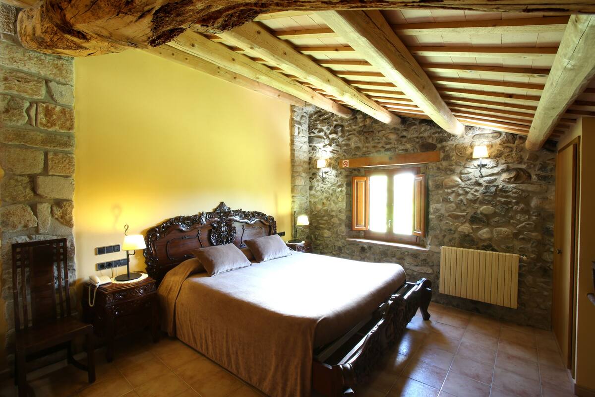 Ein Schlafzimmer mit einer Steinsandsteinwand, Holzbalkendecke, einem Bett mit Kopfteil, Nachttischen und einem Fenster mit Blick.