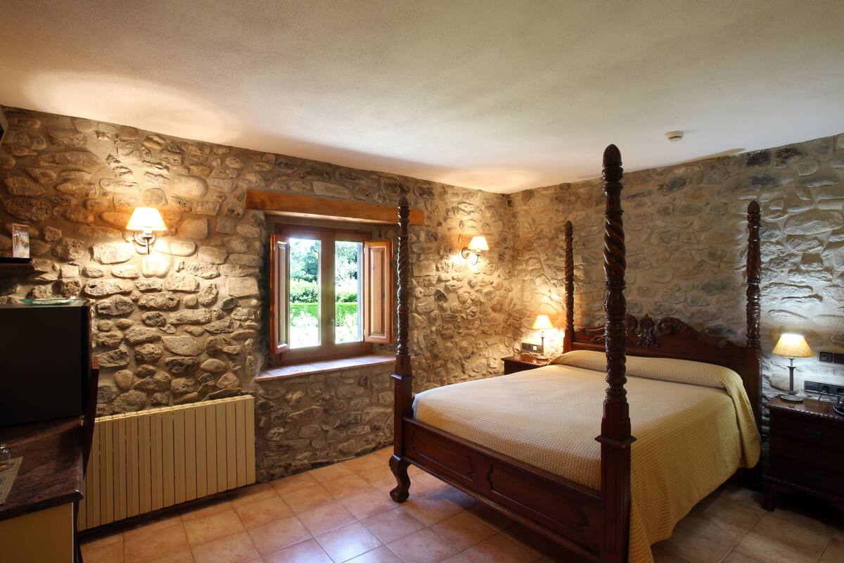 Ein Schlafzimmer mit einer Steinwand, einem Himmelbett, einer Holzkommode und einem Fenster mit Blick auf Grünflächen.