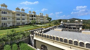 A large, historic building with multiple arched windows and domed structures, surrounded by well-maintained gardens and a clear sky. - The Kumbha Bagh (Kumbhalgarh)