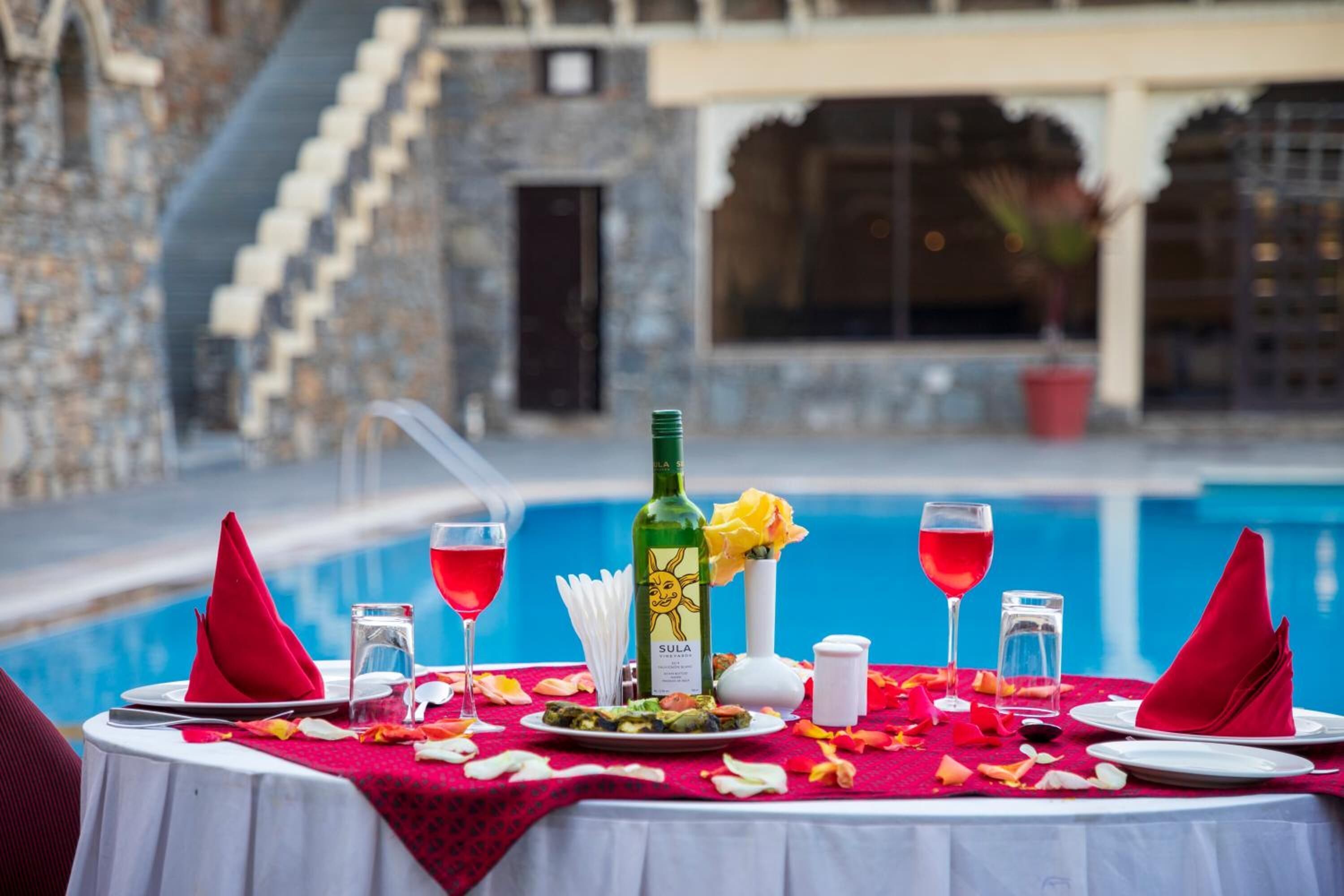 A dining table set for two with a pool view, a bottle of Sula wine, and a red rose.