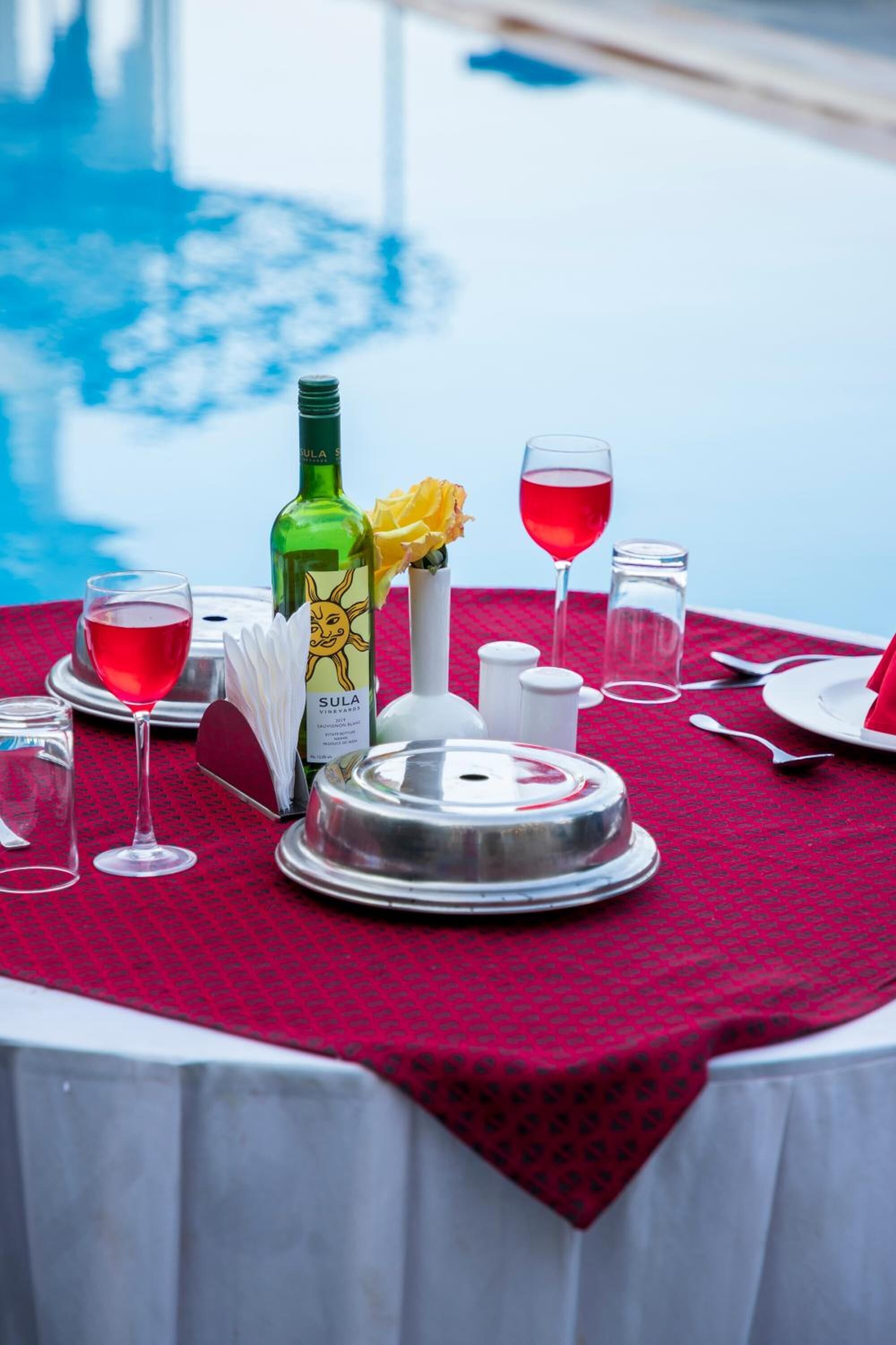 Mesa lista para cenar junto a una piscina, con mantel rojo, una botella de vino Sula, un florero con flores amarillas y saleros y pimenteros.