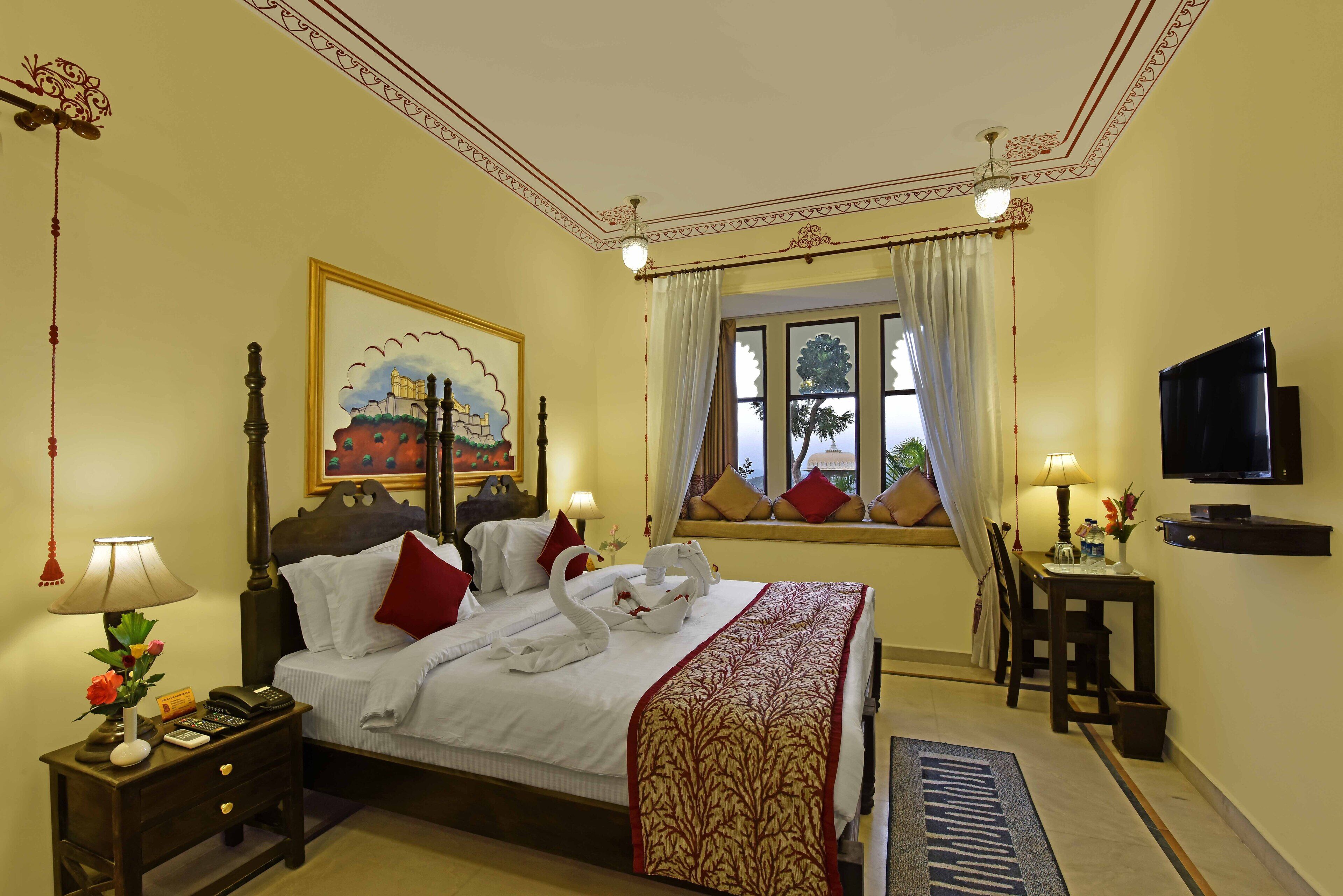 A hotel room with a bed, bedside table, lamp, TV, and a window with curtains.