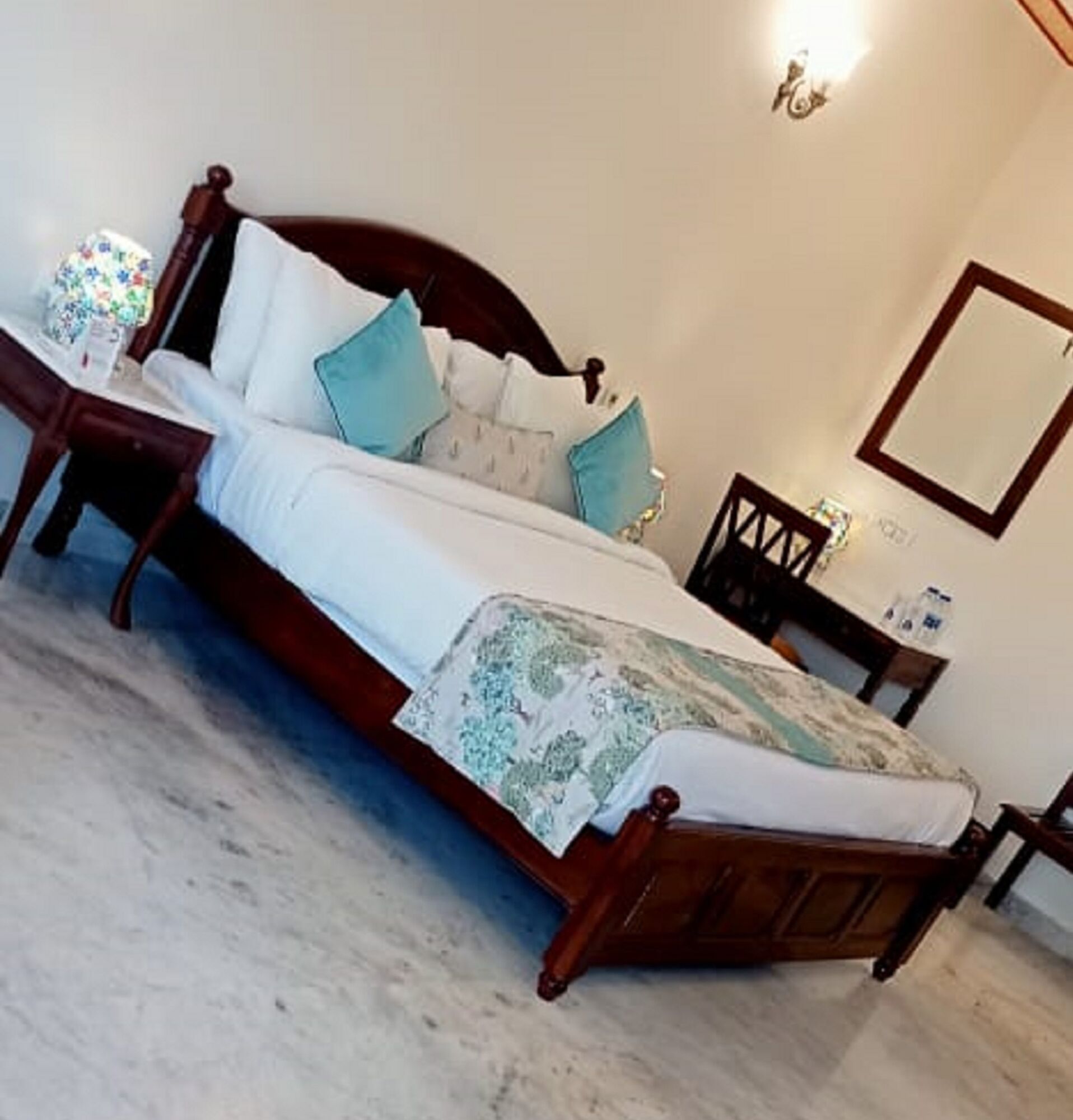 A neatly made bed with a wooden frame, white bedding, and a patterned throw. There is a bedside table with a lamp and a small decorative item. A mirror is mounted on the wall above the bed.