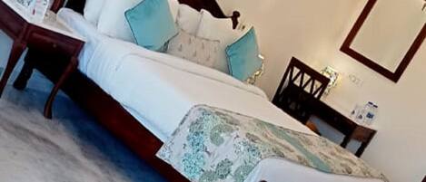 A neatly made bed with a wooden frame, white bedding, and a patterned throw. There is a bedside table with a lamp and a small decorative item. A mirror is mounted on the wall above the bed.