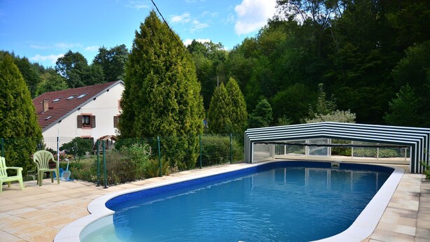 Une piscine avec une terrasse carrelée, entourée de verdure et une maison avec un toit rouge.
