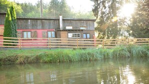Une cabane rustique avec une façade en bois, une petite dépendance attenante et une clôture en bois entourant un étang.