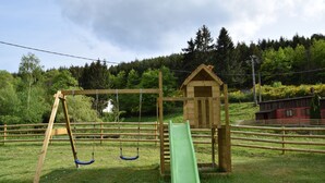 Une aire de jeux avec des balançoires et un toboggan, entourée d’une clôture en bois et d’une petite maison en bois.