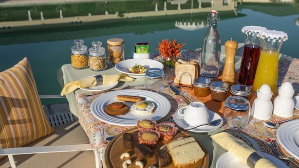 Una mesa preparada para el desayuno con diversas comidas, una botella y un vaso de jugo de naranja junto a una piscina.