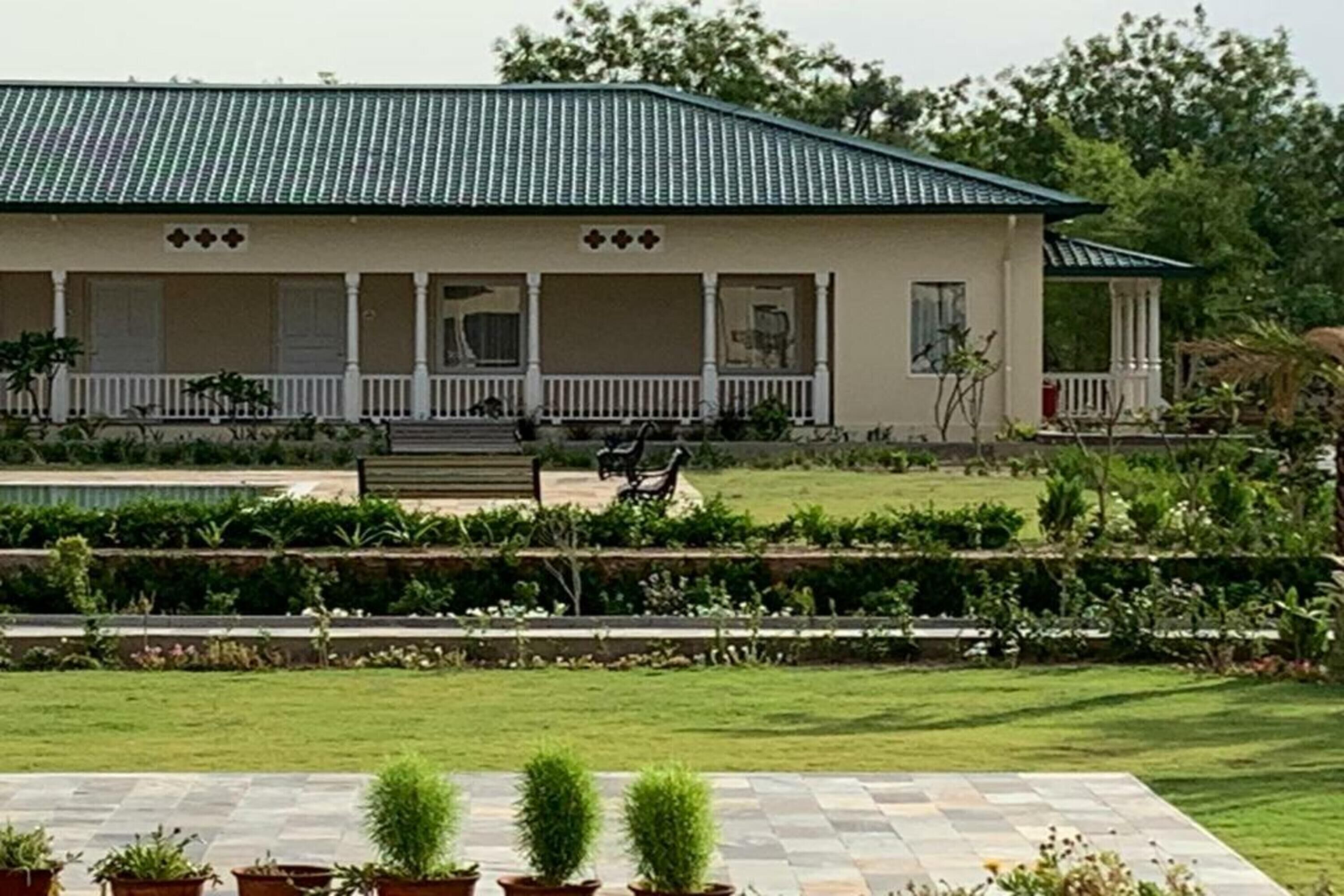 A two-story house with a green roof, a porch with white railings, and a well-maintained garden with various plants and a lawn.