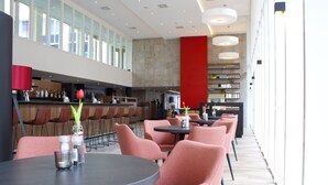 A modern restaurant with round tables and pink chairs, a bar area, and large windows providing natural light. - Bastion Hotel Arnhem (Arnhem)