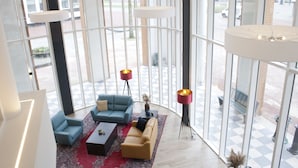 A spacious lobby with a checkered floor, a blue sofa, a beige sofa, and a red rug. There are two tall floor lamps and a large window providing a view of the outside. - Bastion Hotel Arnhem (Arnhem)