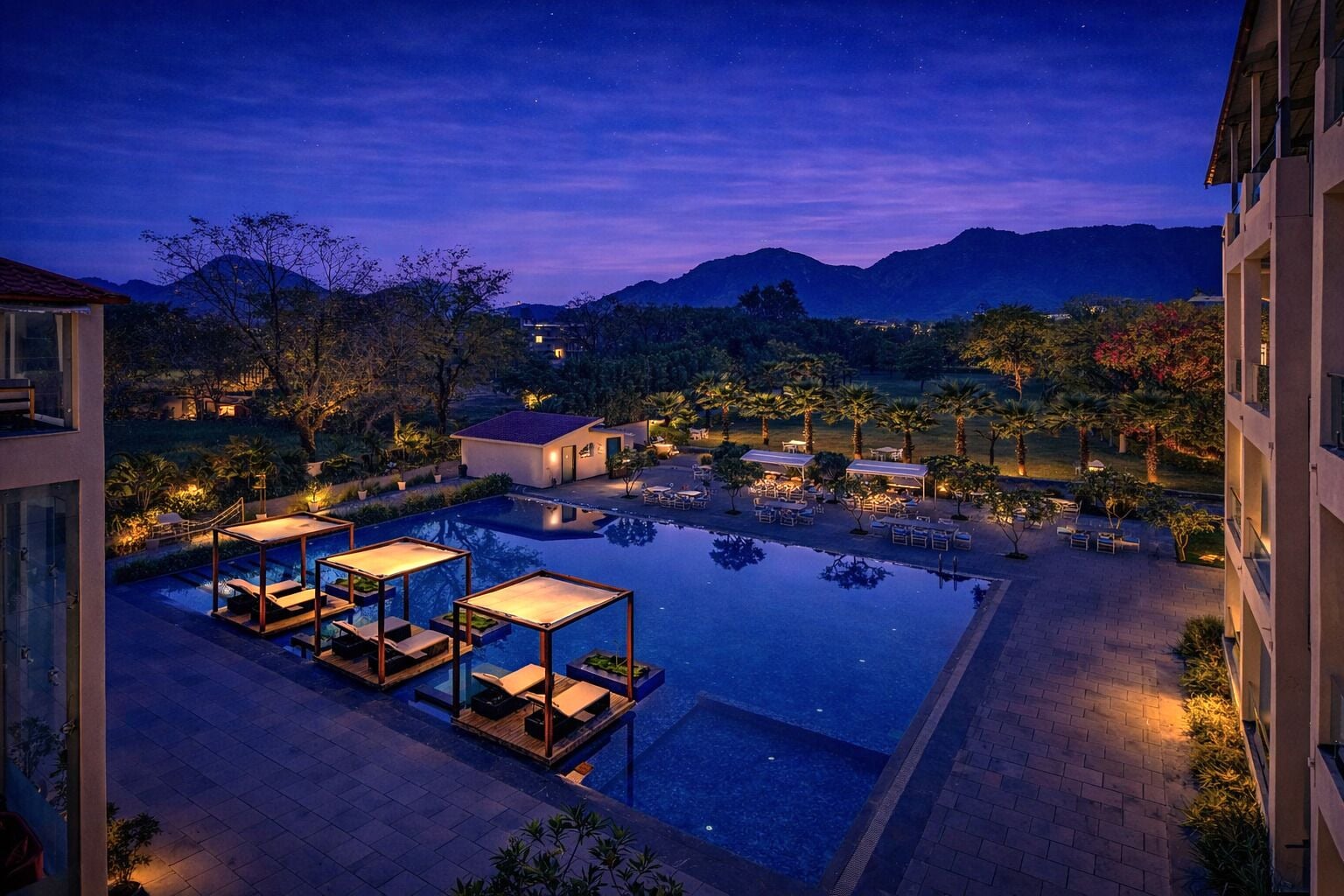 A pool area with lounge chairs and a small building, surrounded by trees and mountains in the background.