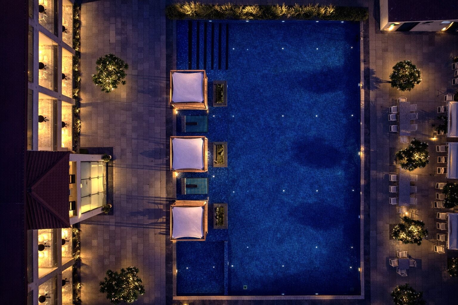 A rooftop pool with four sun loungers and a small table, surrounded by trees and a building with illuminated windows.