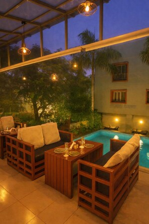 A poolside area with wooden furniture, a glass-enclosed ceiling, and a view of the pool and surrounding greenery. - Hotel Marigold With Swimming Pool By SAH (Abu Road)
