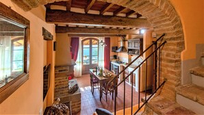 Interior rústico com um arco de pedra, área de jantar com mesa e cadeiras, cozinha com fogão e escada.