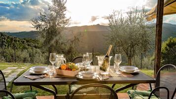 Un tavolo da pranzo all'aperto apparecchiato con piatti, bicchieri e una ciotola di frutta, con vista su un paesaggio montuoso.