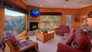 Interior acogedor de una cabaña con chimenea, televisor y un amplio ventanal con vistas a la montaña.