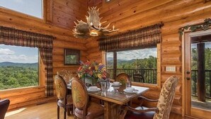 Una cabaña de madera con una mesa de comedor dispuesta para cuatro, rodeada de sillas, una araña y vistas al bosque.