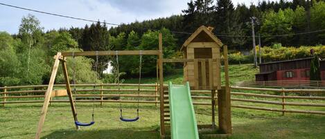 A playground with swings and a slide, surrounded by a wooden fence and a small wooden house.