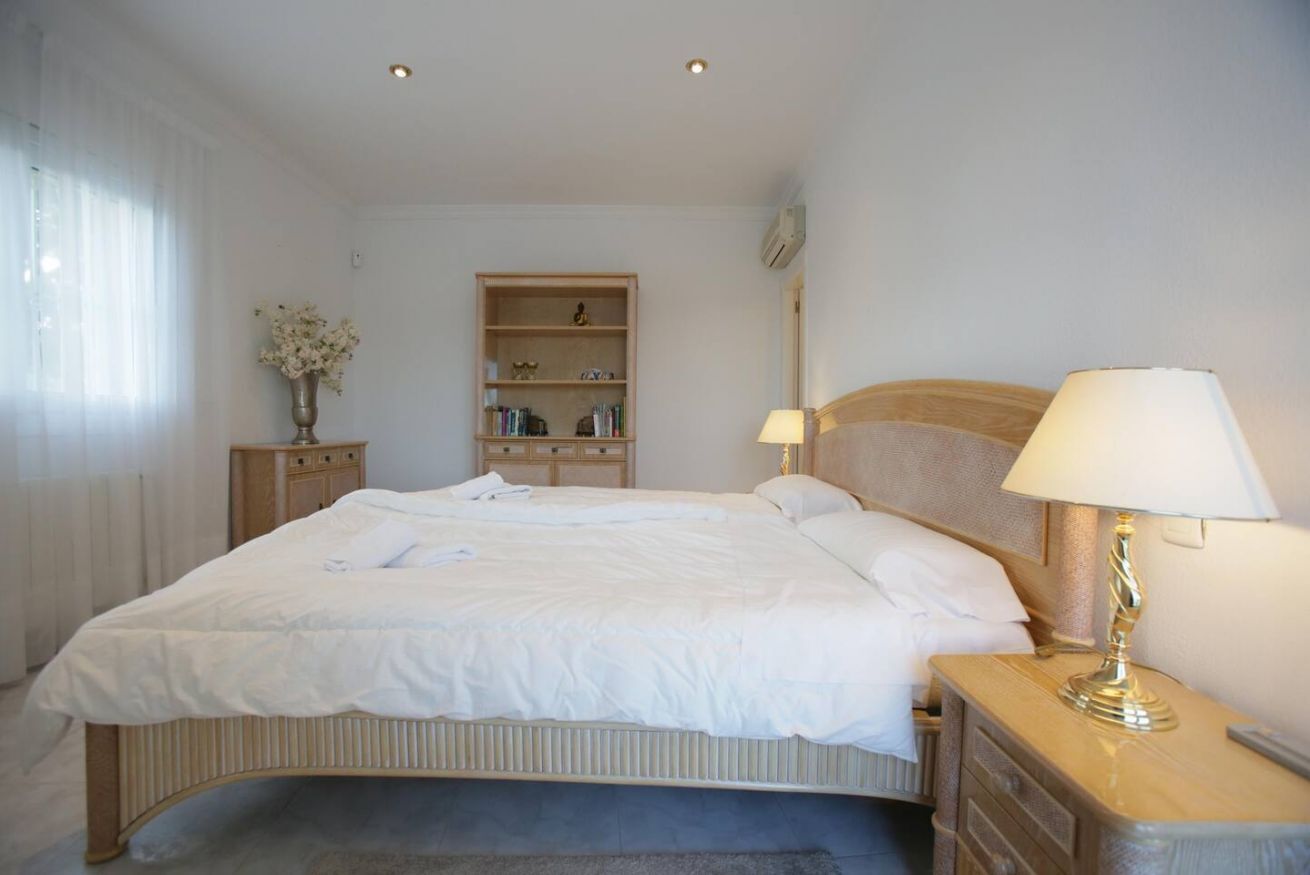 A neatly made bed with white linens, a bedside table with a lamp, a wooden nightstand, a bookshelf, and a radiator.