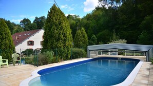 A swimming pool with a tiled deck, surrounded by greenery and a house with a red roof.