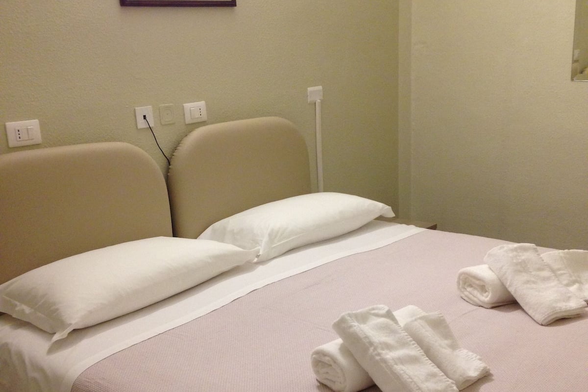 A neatly made bed with pillows and rolled towels, a framed picture on the wall, and a power outlet.