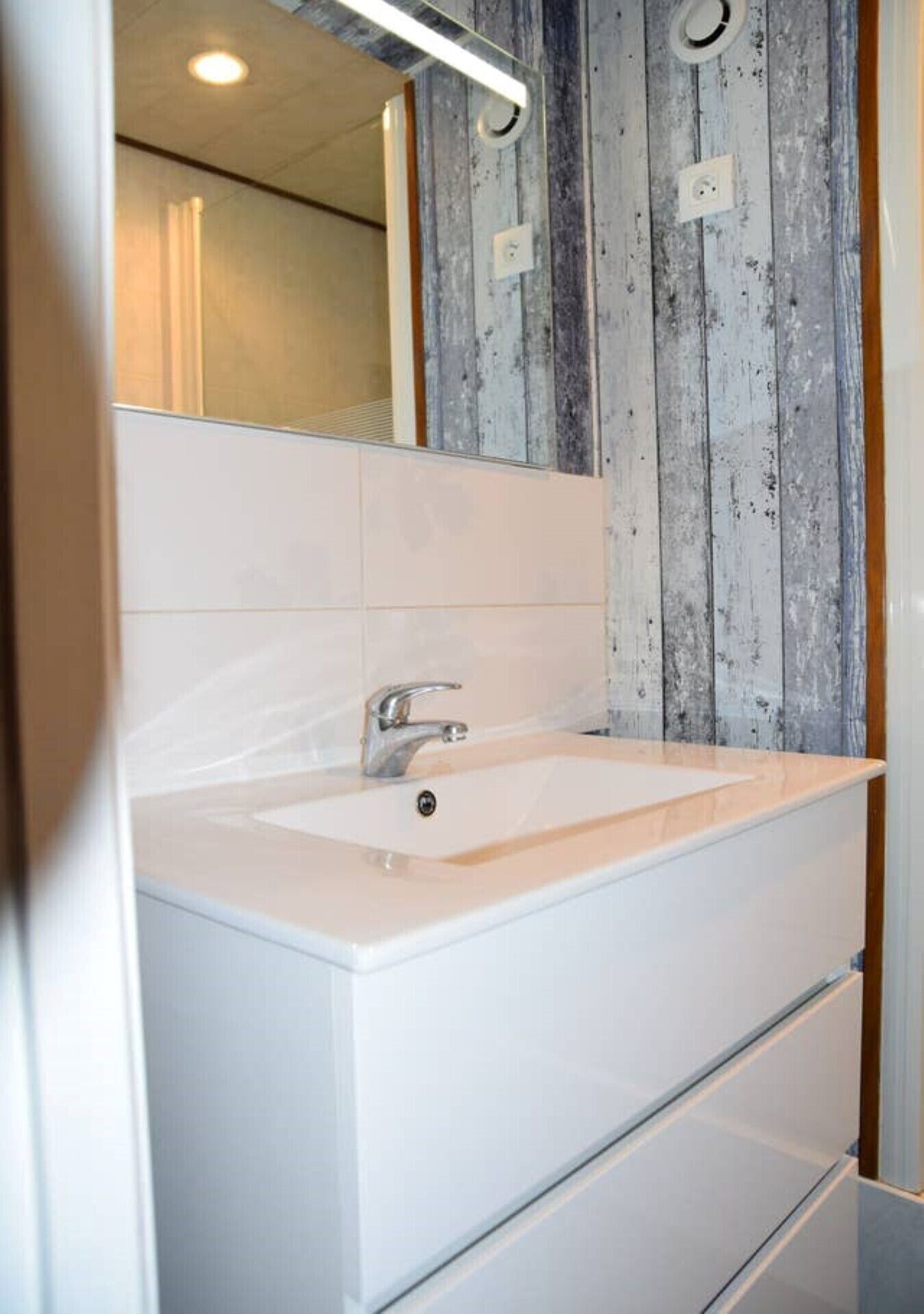 Un baño con lavamanos blanco, un espejo y un panel de pared de madera.