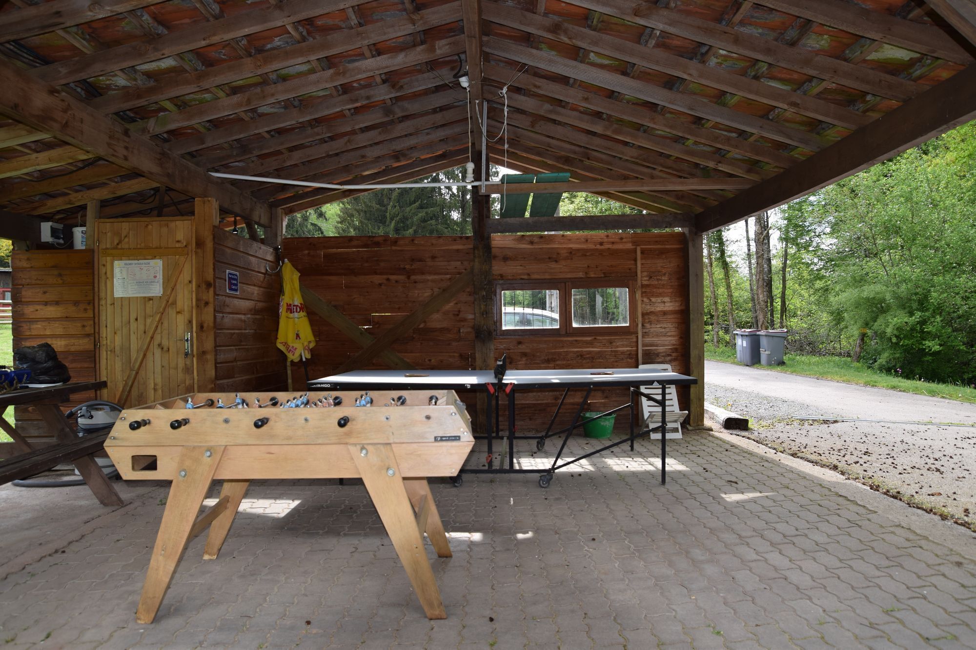 A wooden outdoor seating area with a foosball table, a table with chairs, and a covered section with windows.