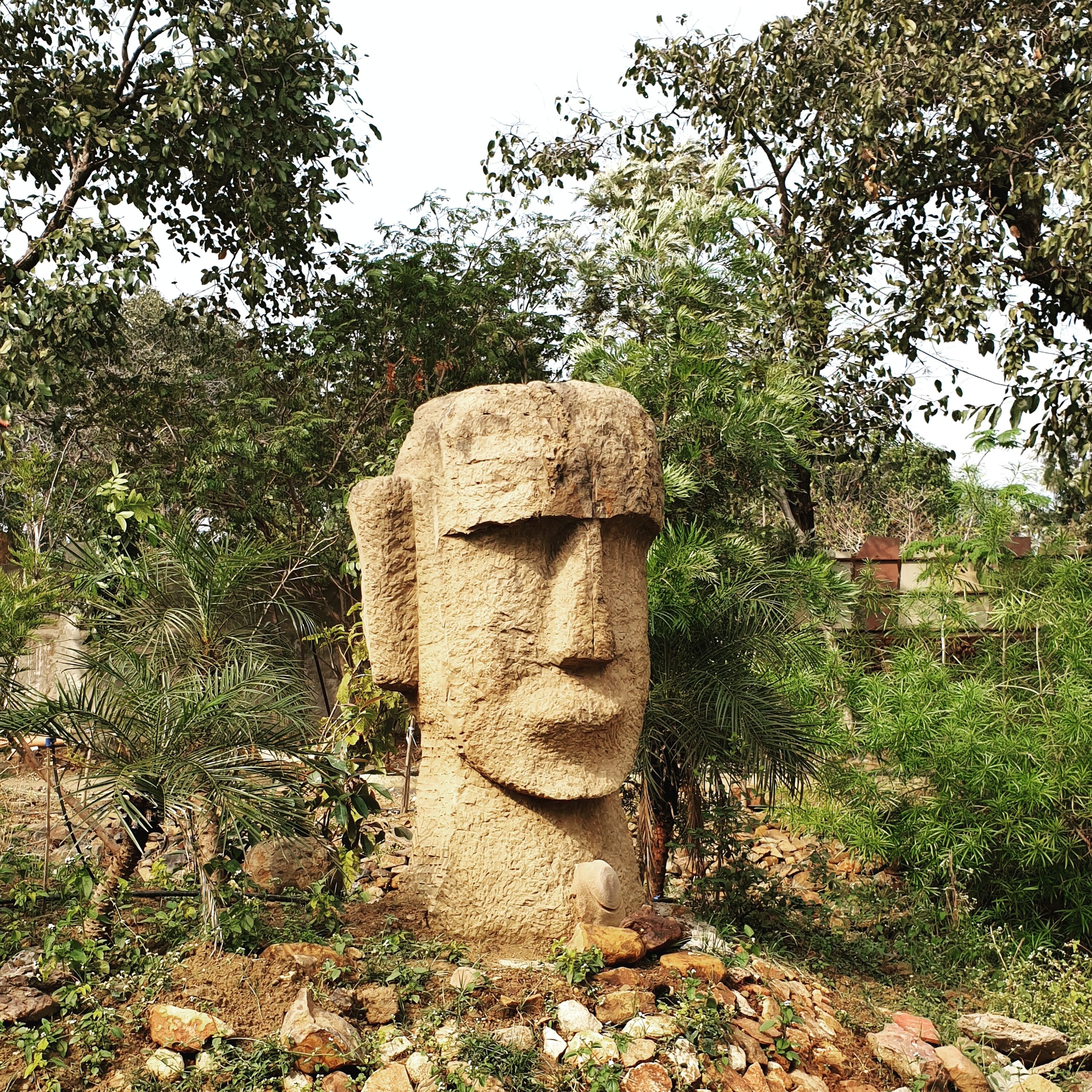 A large stone statue in a garden setting with surrounding vegetation and rocks.