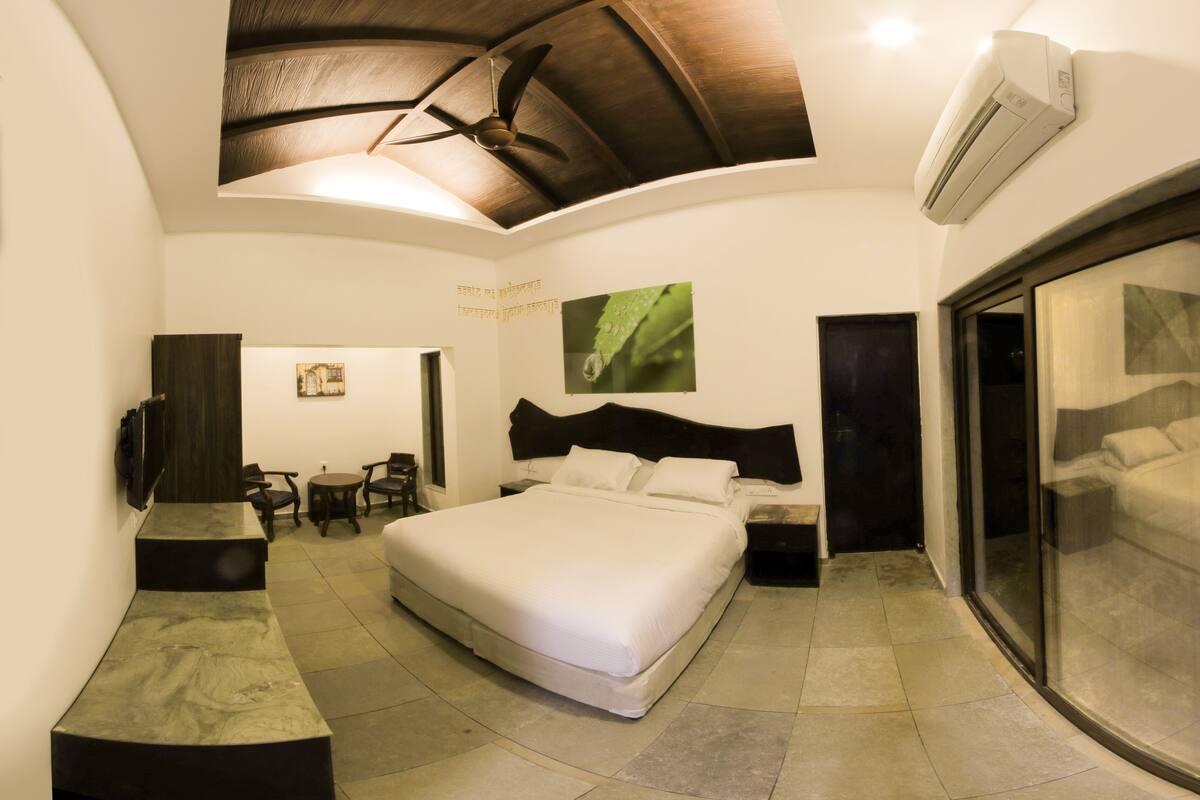 A bedroom with a large bed, a ceiling fan, an air conditioning unit, a desk with chairs, and a large window.