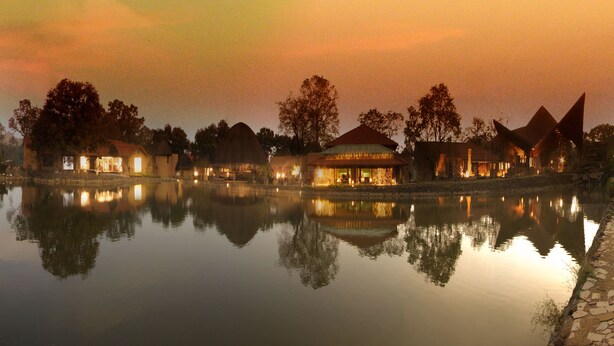 A lakeside hotel with multiple buildings reflecting on the water, featuring a variety of roof styles and a calm evening ambiance. - Serendipity Lakes and Resorts (Tamia)