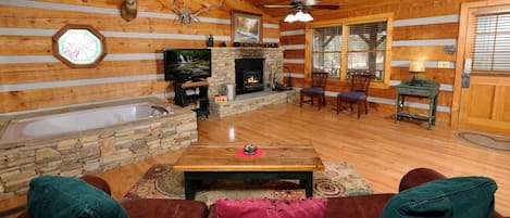 A log cabin interior with a stone fireplace, a stone wall, a wooden ceiling, a ceiling fan, a television, a fireplace mantel, a dining table with chairs, and a leather couch.