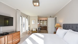 Habitación de hotel con una cama grande, un televisor en un aparador de madera, una mesita con lámpara y vistas a la cocina y al baño a través de una puerta abierta.