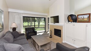 A living room with a sofa, coffee table, fireplace, TV, and a view of a golf course.