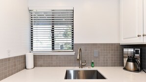 Una cocina con un lavamanos de acero inoxidable, una ventana con persianas, una cafetera y un salpicadero de azulejos.