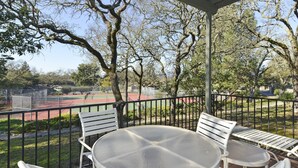Un porche cubierto con una mesa redonda y sillas blancas, con vista a una cancha de tenis y árboles.