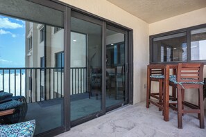 A balcony with a sliding glass door, a wooden table with two chairs, and a patterned cushion. - World Class Sunsets on the Gulf From Your Lanai! Beachfront 2B/2B Sandarac Vacation Condo (Fort Myers Beach)