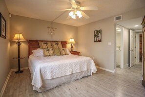 A bedroom with a bed, two bedside lamps, a ceiling fan, and a door leading to another room.