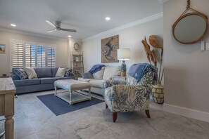 Un salon comprenant un canapé, un fauteuil et un pouf, un ventilateur de plafond et un miroir fixé au mur.