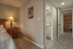 A hotel room with a bed, a nightstand with a lamp, a painting of a palm tree, and a bathroom visible through an open door. - World Class Sunsets on the Gulf From Your Lanai! Beachfront 2B/2B Sandarac Vacation Condo (Fort Myers Beach)