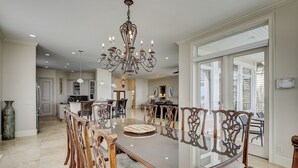A spacious dining area with a large glass table surrounded by ornate chairs, a chandelier, and a view of the kitchen and living areas. - 81 Dune Lane - Oceanfront 6BR | Private Pool & Spa | Sleeps 18 (Hilton Head Island)