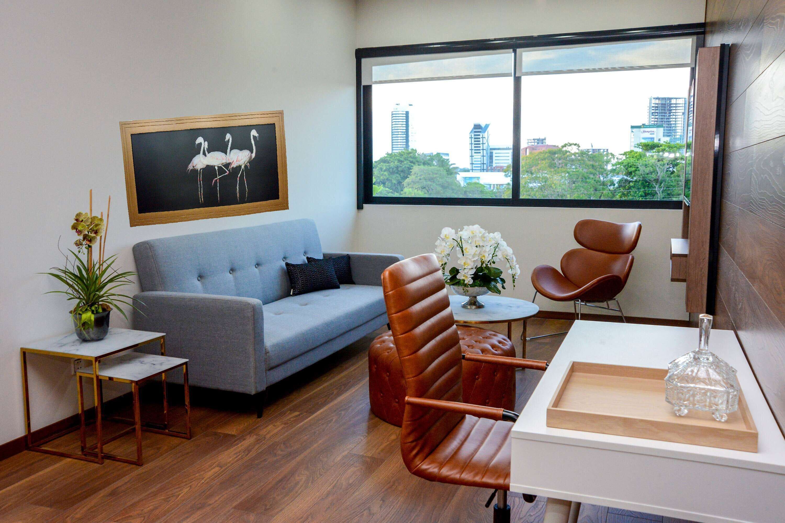 A modern living room with a grey sofa, a brown leather chair, a small table with a vase, and a view of the city through a large window.