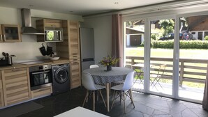 Une cuisine moderne avec des meubles en bois, un four encastré et un lave-linge. On y trouve un coin repas avec une table et des chaises, ainsi qu’une grande porte-fenêtre coulissante donnant sur un patio extérieur.