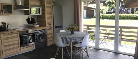 Una cocina moderna con armarios de madera, horno integrado y lavadora. Hay un comedor con mesa y sillas, y un amplio ventanal corredera que da a un patio exterior.