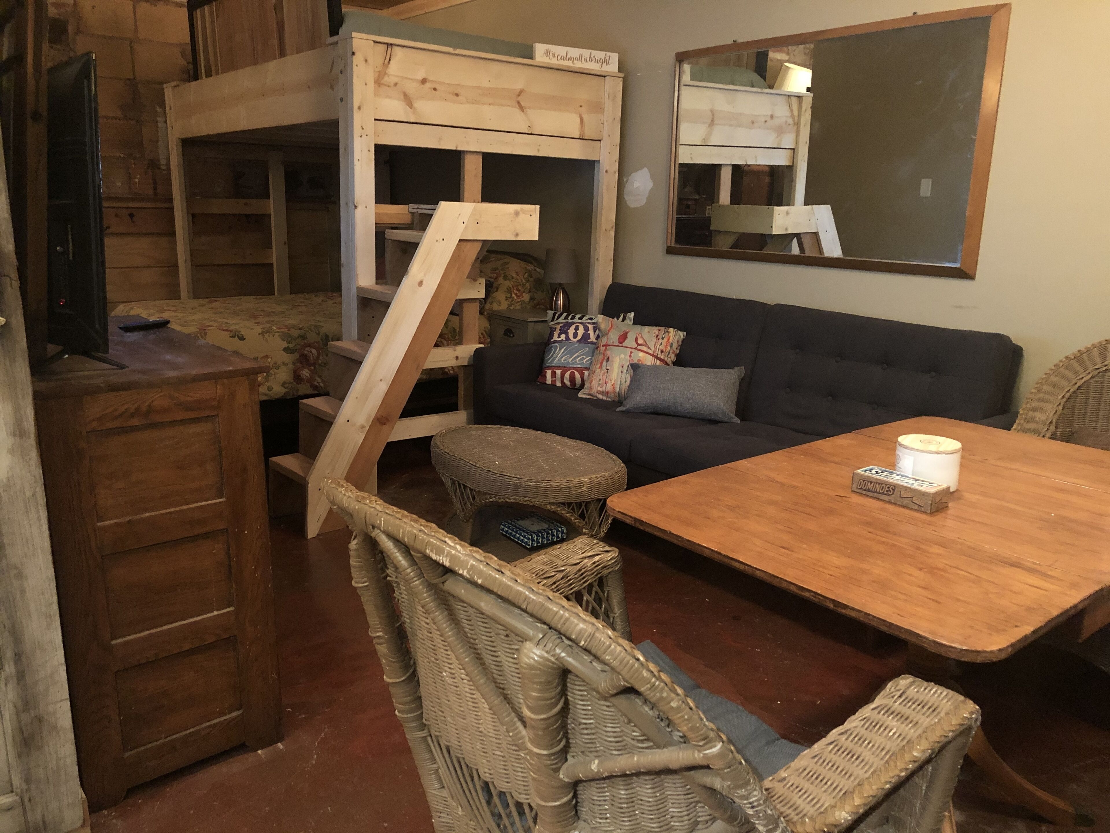 A cozy room with a wooden bunk bed, a dark sofa, a wicker chair, and a small table with a box of dominoes.
