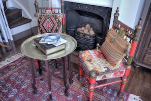 A traditional room with a patterned rug, a wooden chair with a colorful cushion, a small round table with books, and a fireplace with logs.